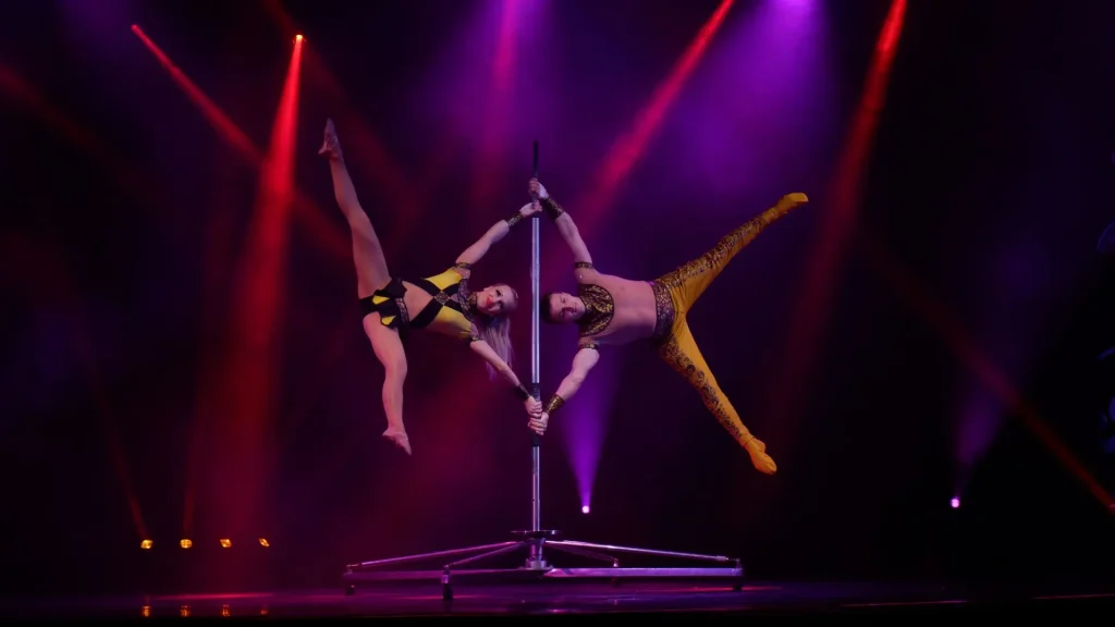 Two acrobatic artists holding a dramatic pose on a free-standing pole under vibrant purple and red stage lights.